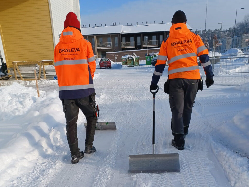 Kaksi lumitöitä tekevää työntekijää kävelee lumisella pihalla lumentyöntimet kädessään; heillä on kirkkaanoranssit huomiotakit, joissa lukee BRALEVA, ja taustalla näkyy rakennuksia.