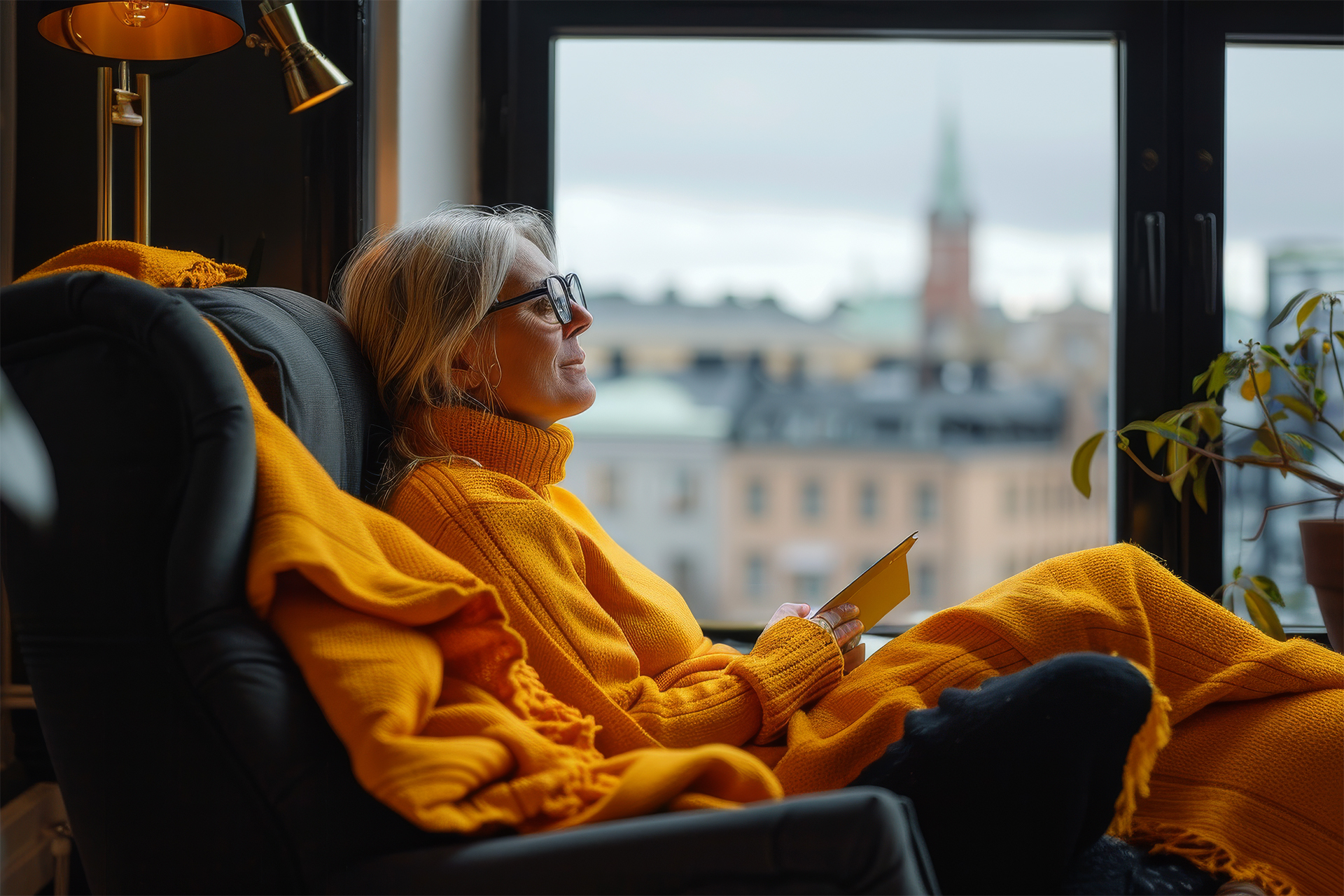 Oranssipaitainen silmälasipäinen nainen istuu ikkunan edessä ja hymyilee. Kädessään hänellä on kirja auki.