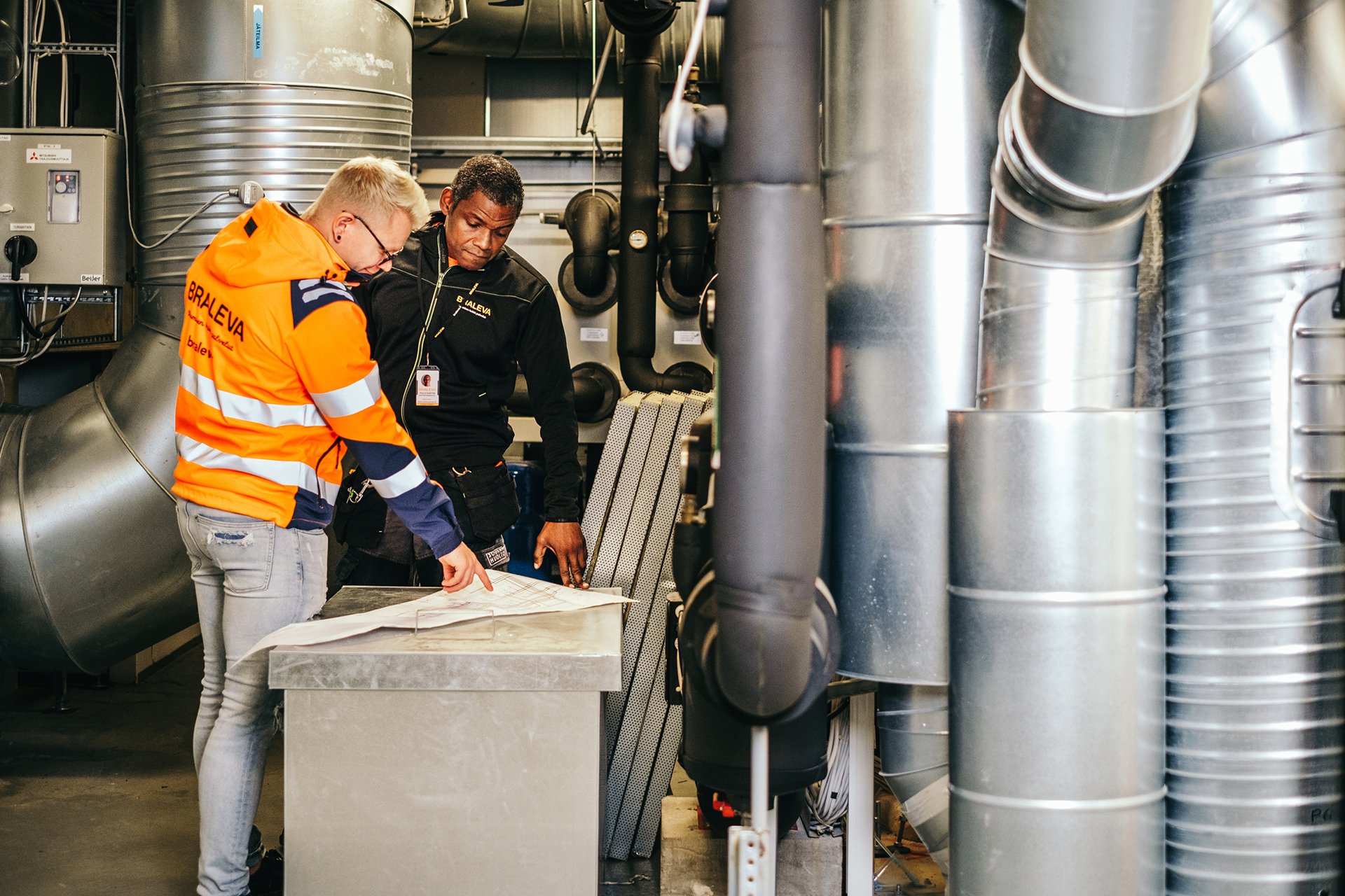 Kaksi miestä Bralevan työvaatteissa katsoo rakennuspiirustuspaperia, joka on harmaalla pöydällä. Ympärillä ilmastointiputkia.