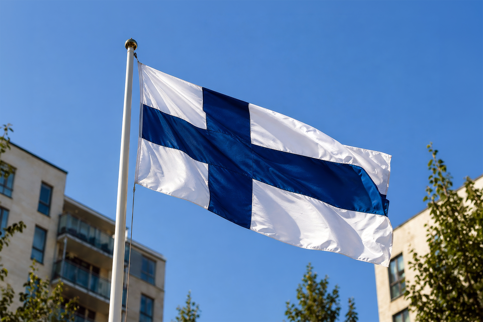 Suomen lippu lipputangossa, kesäinen aurinkoinen valo, taustalla kerrostaloja ja puita.